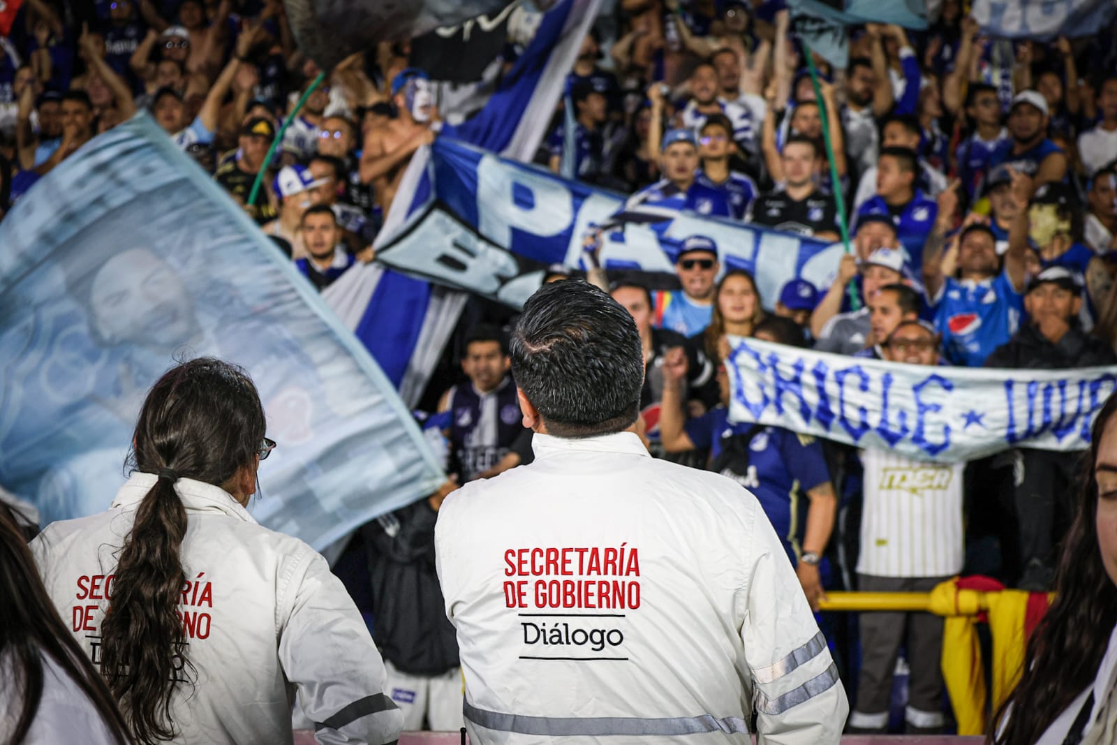 Foto donde se ven dos funcionarios de Diálogo de la SDG mirando hacia la tribuna de los hinchas de Millonarios que están alentando a su equipo.