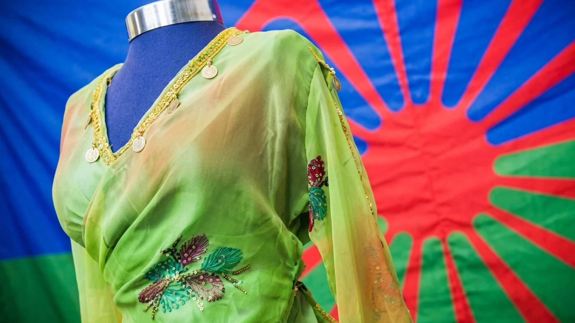 Foto de un vestido de mujer caracteristico del pueblo Rrom y de fondo la bandera del pueblo Rrom