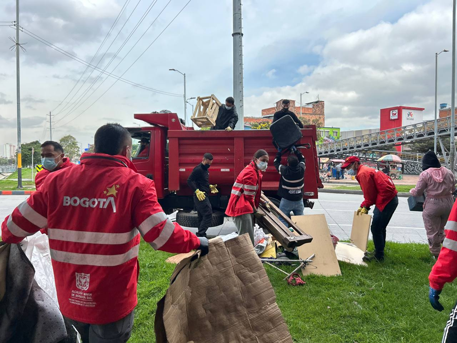 Se observa un camión rojo donde varias personas con chaquetas institucionales de Bogotá cargan muebles, madera y otros elementos retirados de la zona verde junto a la avenida. Al fondo se aprecia un puente peatonal y edificaciones del sector.
