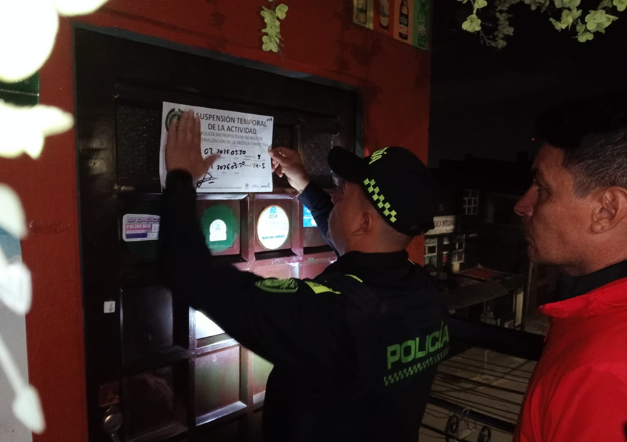 Un oficial de la Policía Nacional pega un cartel de "Suspensión Temporal de la Actividad" sobre la puerta de madera de un establecimiento comercial en Puente Aranda. A su lado, un funcionario con chaqueta roja supervisa la acción. La imagen captura uno de los cuatro cierres realizados durante un operativo nocturno de inspección y vigilancia en Bogotá para garantizar el cumplimiento de normas sanitarias y de seguridad.