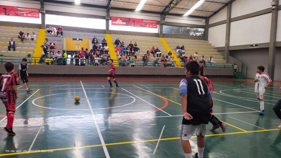 Niños jugando fútbol en una cancha cubierta de coliseo. En primer plano, un jugador con camiseta negra número 11 observa el balón en el centro de la cancha, mientras otros niños con uniformes rojo y negro se mueven hacia la jugada. En las gradas amarillas del fondo hay varias personas sentadas viendo el partido.