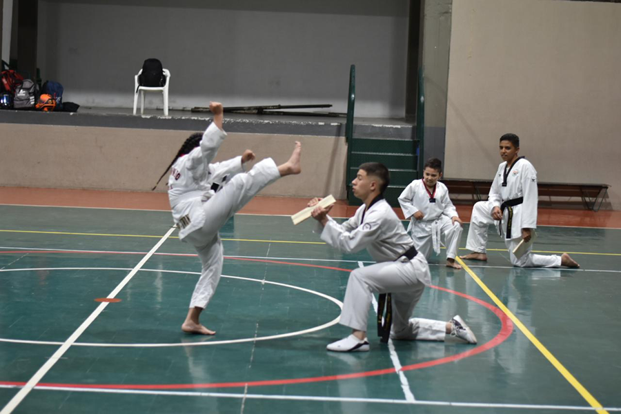 Niña practicando taekwondo realiza una patada alta en una cancha cubierta, mientras un compañero arrodillado sostiene una tabla de madera para que la rompa. Al fondo, otros niños con uniforme blanco observan la demostración desde el borde de la cancha.