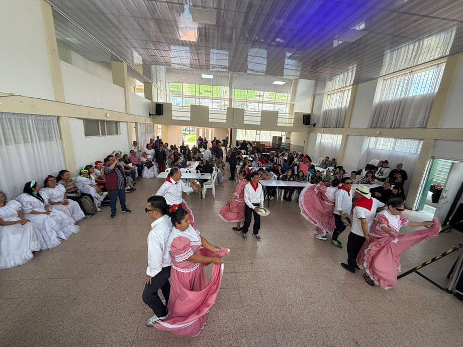 Grupo de parejas bailando una danza folclórica en un salón comunitario amplio y bien iluminado. Las mujeres visten vestidos rosados con vuelos y los hombres camisas blancas con pañuelo rojo al cuello. Alrededor, numerosas personas están sentadas en mesas y sillas observando la presentación.