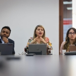 En la foto se ve una mujer dirigiéndole la palabra a la mesa, a su derecha un hombre afrocolombiano y a su izquierda una mujer con gafas.
