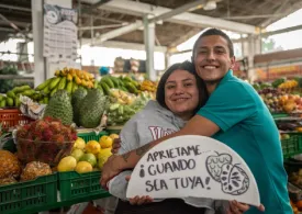 Una pareja de jóvenes mujer y hombre abrazándose, sonriendo cargan un aviso que dice "Aprietame... ¡cuando sea tuya!"  y de fondo un puesto de frutas de la plaza de mercado