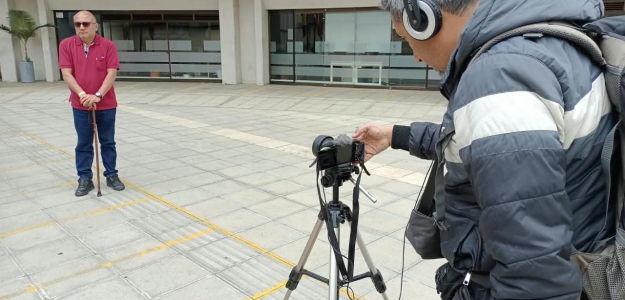 Foto de dos personas, una con una cámara grabando a un hombre que se encuentra de pie mirando a la cámara