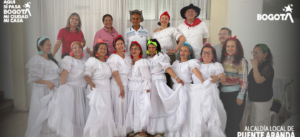Grupo de personas adultas y adultas mayores posando en un salón comunitario. Varias mujeres visten trajes blancos tradicionales con faldas amplias y detalles en encaje, acompañadas de accesorios coloridos en el cabello; junto a ellas hay hombres y otras personas con vestimenta casual. 