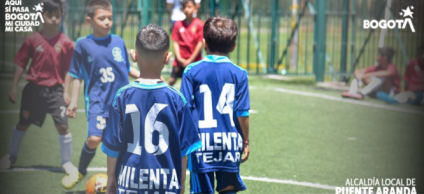 Niños jugando fútbol en una cancha sintética cerrada con malla metálica. En primer plano, dos niños con uniformes azules y números 16 y 14 en la espalda observan el balón naranja en el suelo, mientras otros niños con camisetas rojas y azules se preparan para la jugada al fondo.