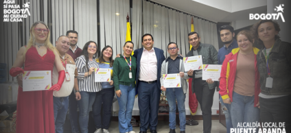 “Grupo de personas posando en un salón de la Alcaldía Local de Puente Aranda luego del acto de posesión del Consejo Local LGBTI 2026–2027. Varias de ellas sostienen certificados en las manos. En el centro se encuentra un funcionario de la Alcaldía acompañado por integrantes electos del Consejo. Al fondo se observan las banderas de Bogotá y de Colombia, y en las esquinas superiores aparecen los logos institucionales de ‘Bogotá’ y de la Alcaldía Local de Puente Aranda.”