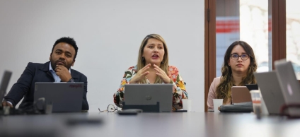 En la foto se ve una mujer dirigiéndole la palabra a la mesa, a su derecha un hombre afrocolombiano y a su izquierda una mujer con gafas.