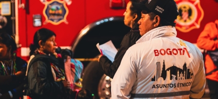 De fondo un carro de bomberos de Bogotá, se encuentra una mujer emebra haciendo fila y en frente dos funcionarios, uno de ellos de chaqueta blanca con el logo de Bogotá y el texto Diálogo