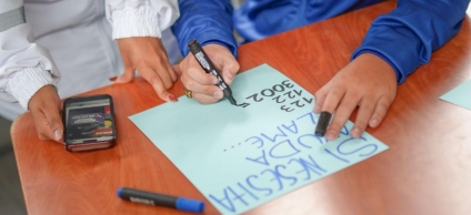 Foto donde se aprecian las manos de un menor de edad escribiendo nñumeros sobre una cartulina y a su lado se aprecian las manos de una persona adulta.