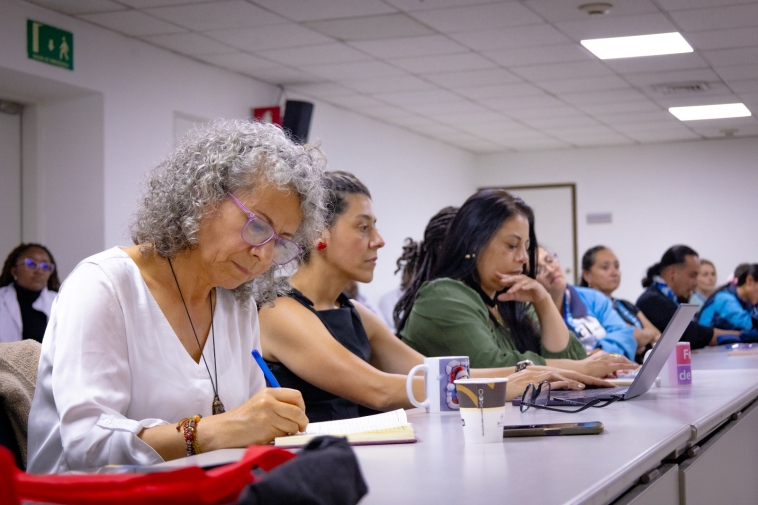 Foto de mujeres en una mesa, se destaca una persona mayos escribiendo sobre una mesa.