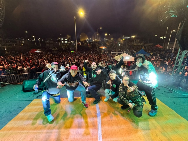 Artistas posan sobre el escenario frente a una multitud durante un festival de hip hop nocturno, en un ambiente lleno de energía, música y cultura urbana.