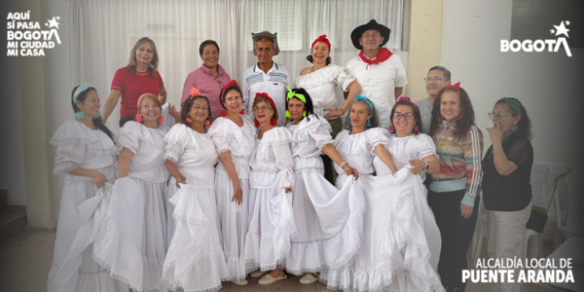 Grupo de personas adultas y adultas mayores posando en un salón comunitario. Varias mujeres visten trajes blancos tradicionales con faldas amplias y detalles en encaje, acompañadas de accesorios coloridos en el cabello; junto a ellas hay hombres y otras personas con vestimenta casual. 