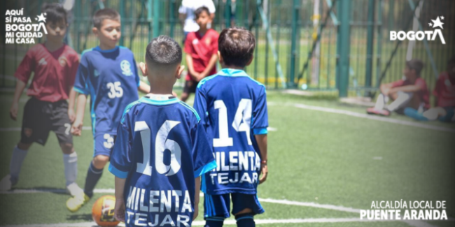 Niños jugando fútbol en una cancha sintética cerrada con malla metálica. En primer plano, dos niños con uniformes azules y números 16 y 14 en la espalda observan el balón naranja en el suelo, mientras otros niños con camisetas rojas y azules se preparan para la jugada al fondo.