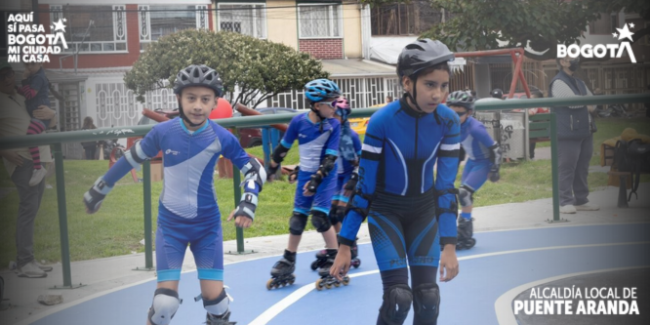 Niñas y niños practicando patinaje en una pista azul al aire libre mientras usan cascos y equipo de protección. Al fondo se observan viviendas, zonas verdes y personas acompañantes. La imagen muestra un ambiente deportivo y comunitario durante una jornada impulsada por la Alcaldía Local de Puente Aranda.