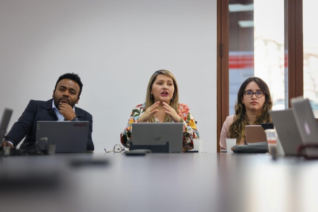 En la foto se ve una mujer dirigiéndole la palabra a la mesa, a su derecha un hombre afrocolombiano y a su izquierda una mujer con gafas.