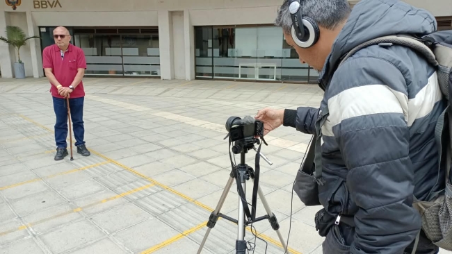 Foto de dos personas, una con una cámara grabando a un hombre que se encuentra de pie mirando a la cámara